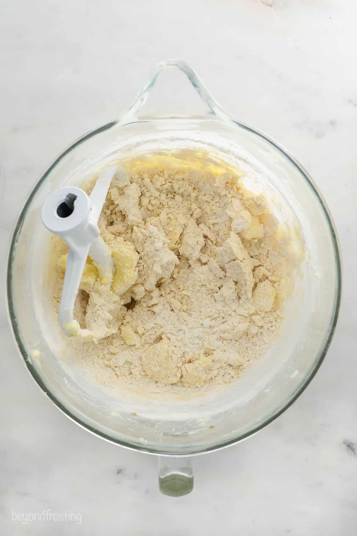 The in-progress cookie dough in a large glass beaker after the dry ingredients have been poured in