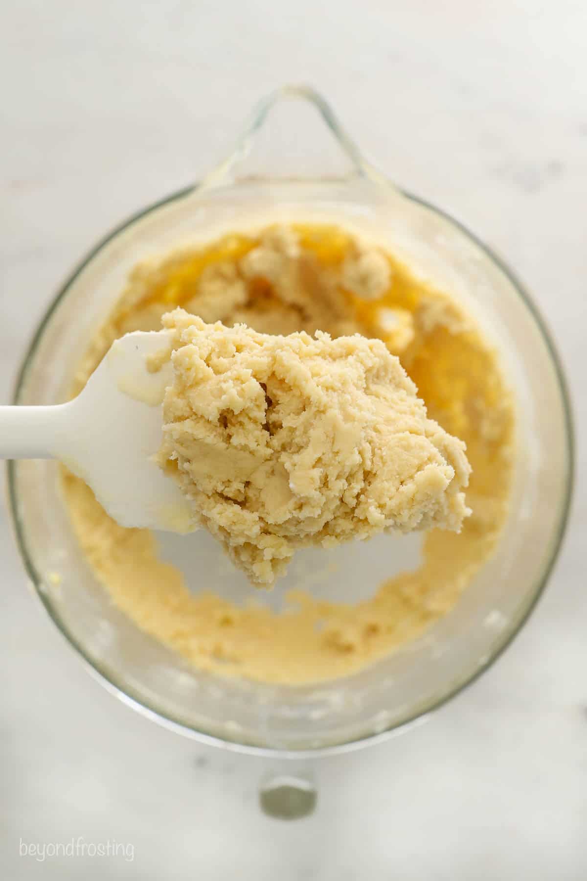 A scoop of cookie dough on a dessert spatula with more cookie dough in a glass beaker underneath it