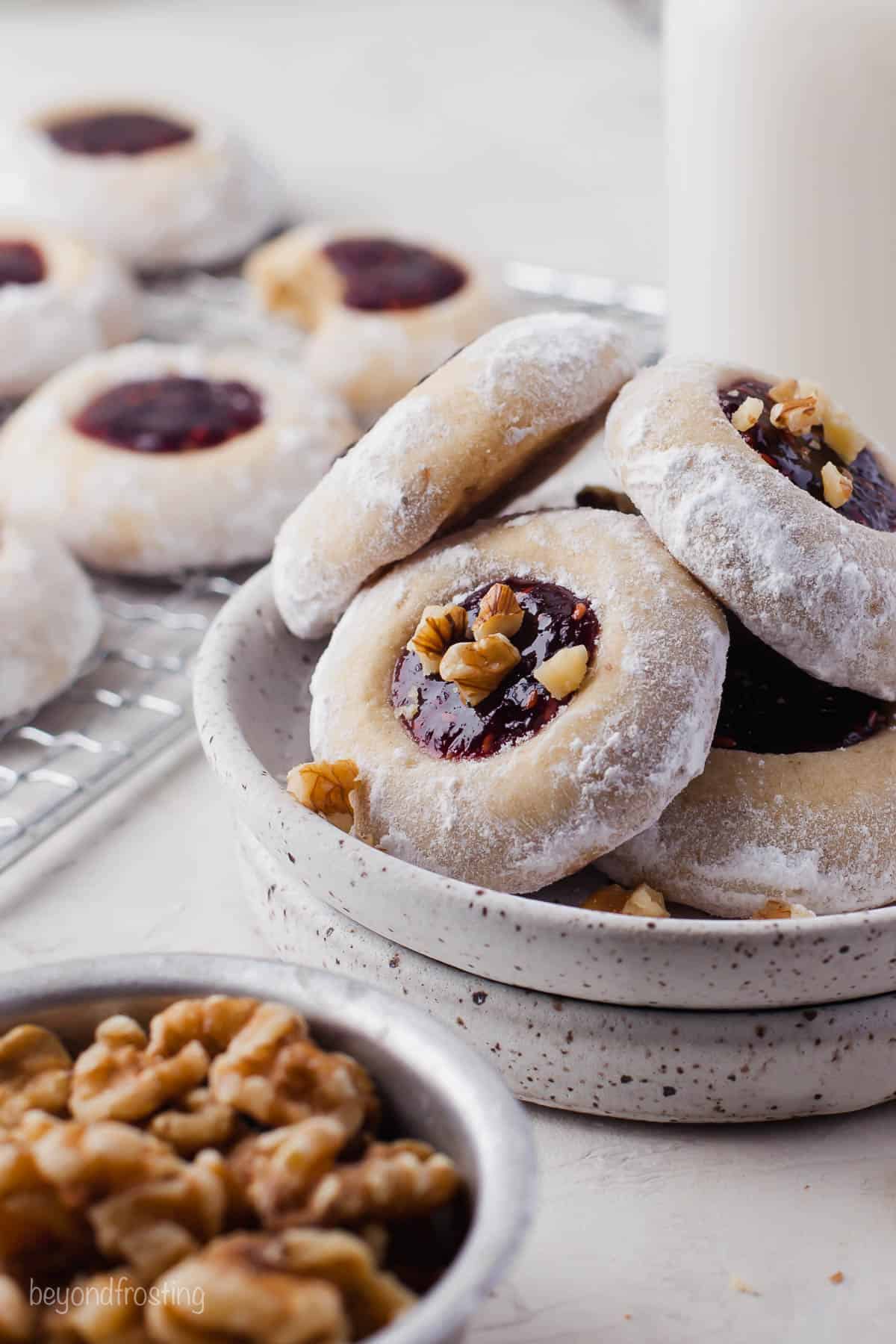 Jam-filled thumbprint cookies piled into a plate with a bowl of walnuts in the foreground