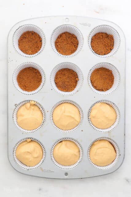 Overhead view of a cupcake pan, 6 liners filled with cookie crumbs and 6 liners filled with pumpkin cheesecake batter