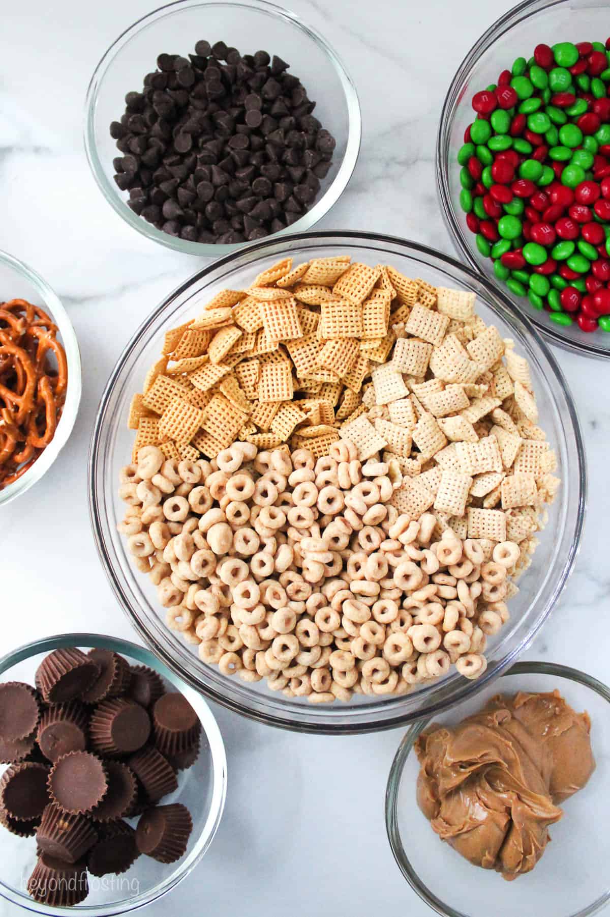 assorted glass bowl with the ingredients for Reindeer Chow Snack Mix like pretzels, chocolate chips, Christmas M&Ms and Reese's Peanut Butter Cups.