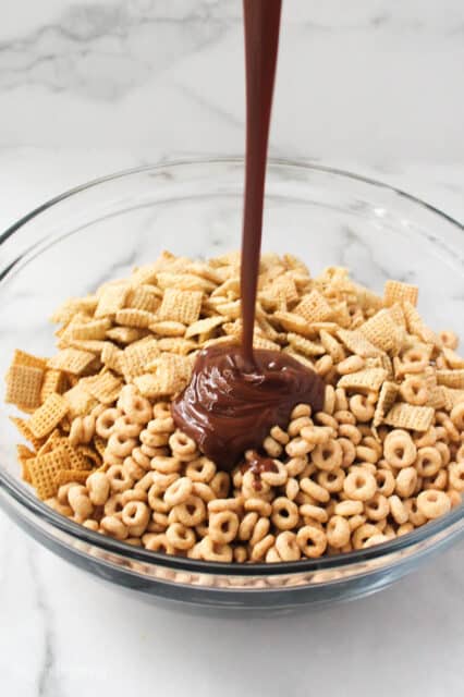 a white glass bowl with Chex cereal, Honey Nut Cheerios, and chocolate pouring over the bowl