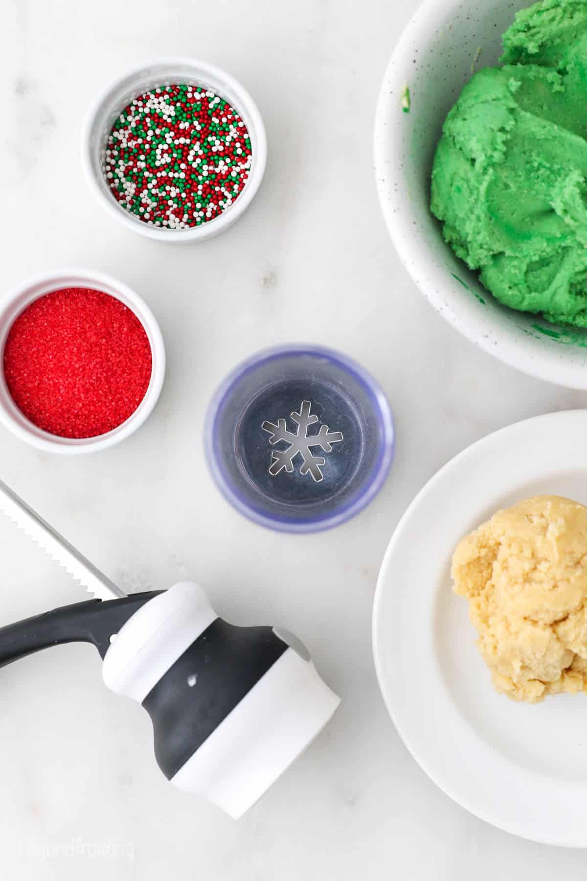 Two dishes of Christmas-themed sprinkles on a counter with spritz cookie dough and a cookie press