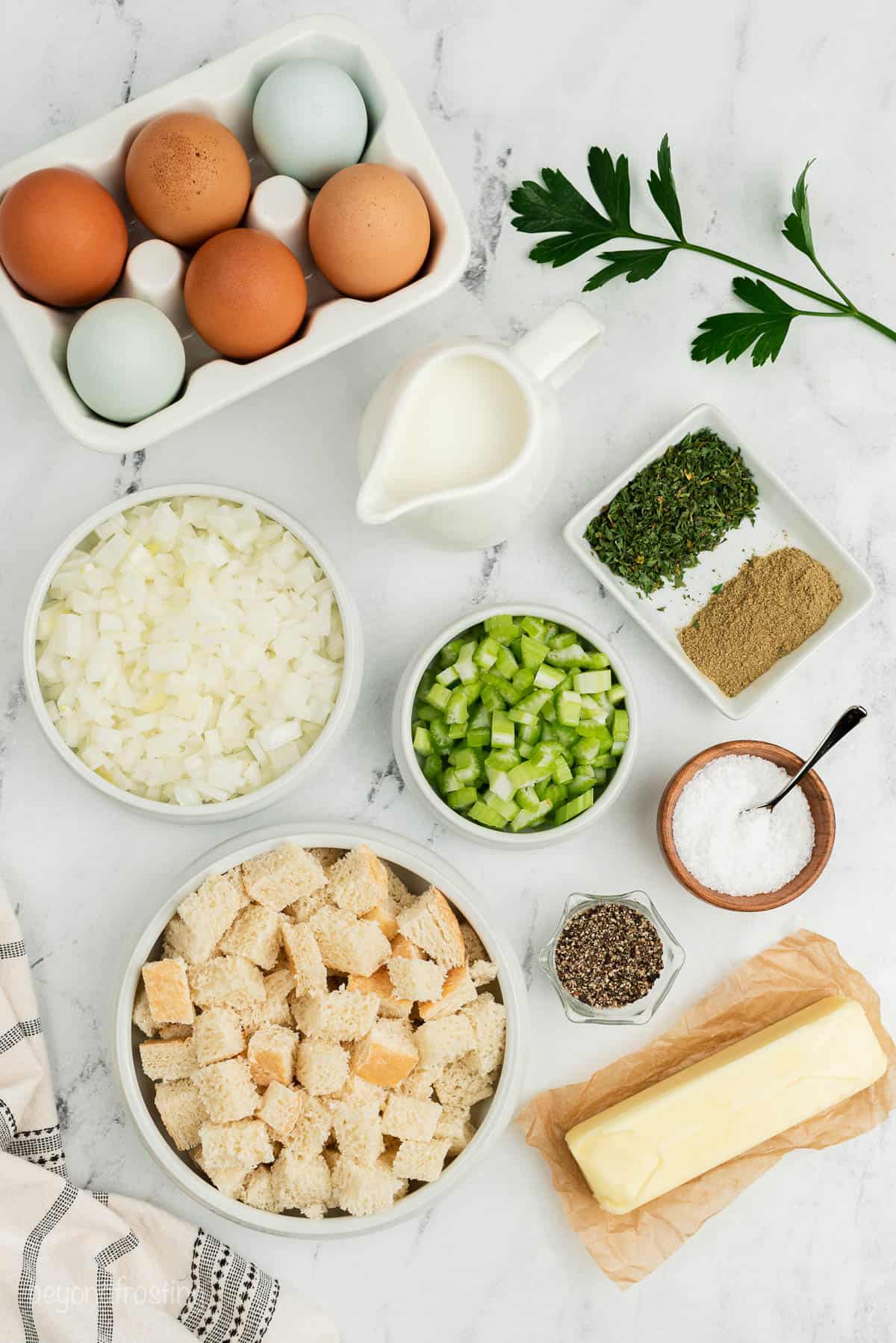 Milk, eggs, butter, salt, pepper, bread cubes and the rest of the stuffing ingredients arranged on a white marble countertop