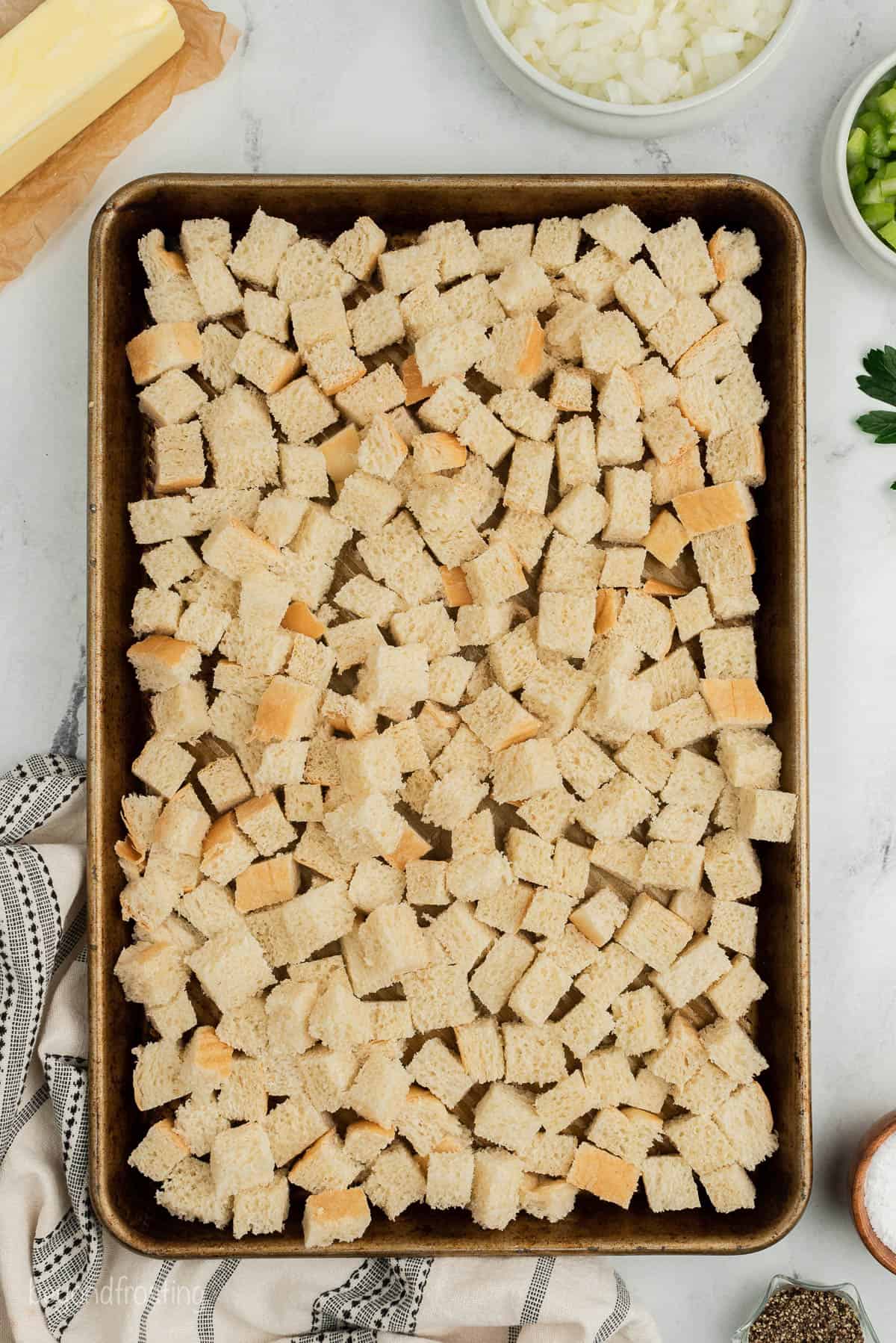 A baking sheet full of sourdough bread cubes on top of a counter with a kitchen towel, a stick of butter and some chopped veggies