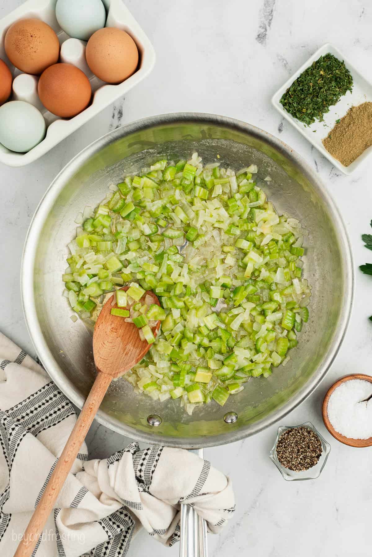 Chopped celery and onions being stirred with a wooden spoon on a countertop with eggs, spices and dried herbs
