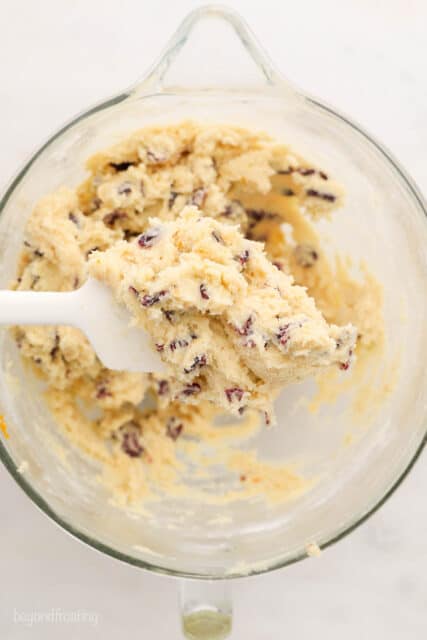 A white spatula with cookie dough with cranberries being held over a glass mixing bowl