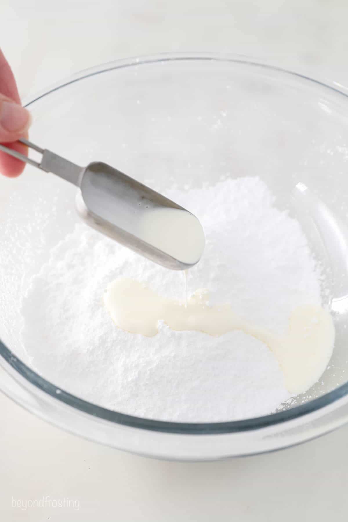Two tablespoons of milk being poured into a bowl full of sifted powdered sugar