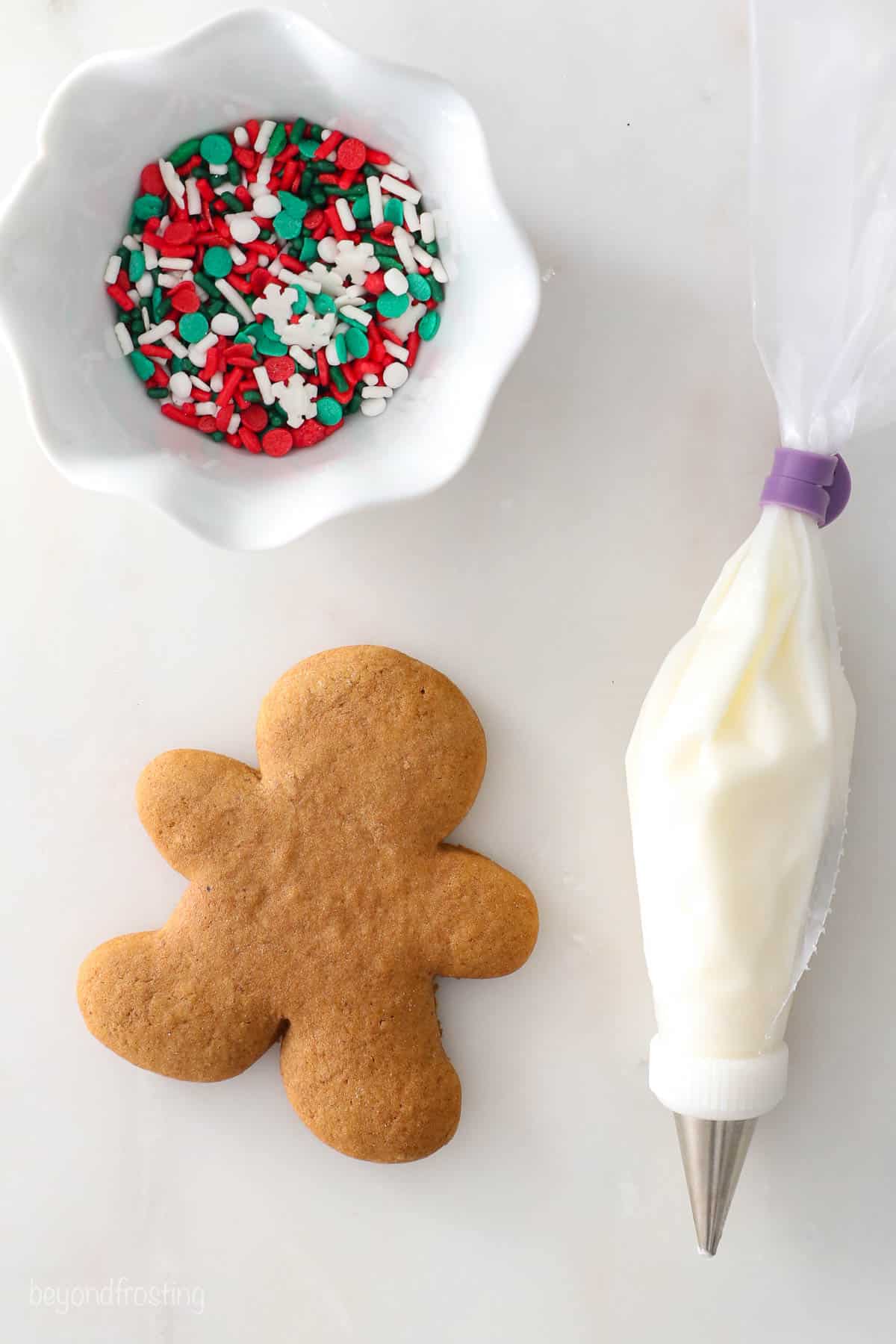 An unfrosted gingerbread cookie beside a bowl of sprinkles and a piping bag filled with icing