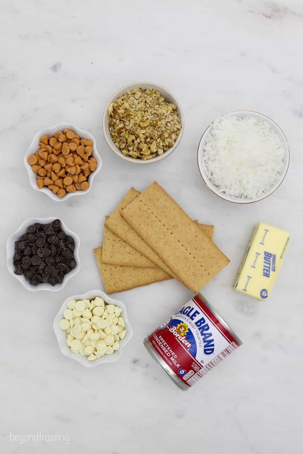 Butterscotch chips, white chocolate chips, sweetened coconut flakes and the rest of the cookie bar ingredients on a countertop