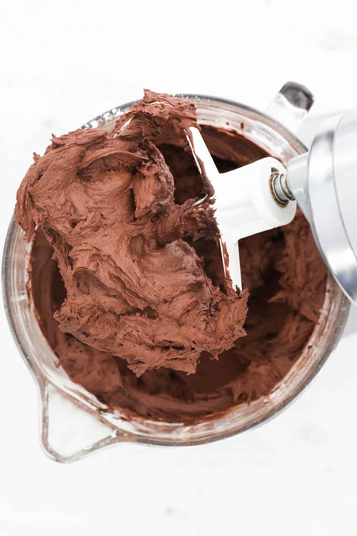 A stand mixer blade of chocolate frosting over a glass mixing bowl