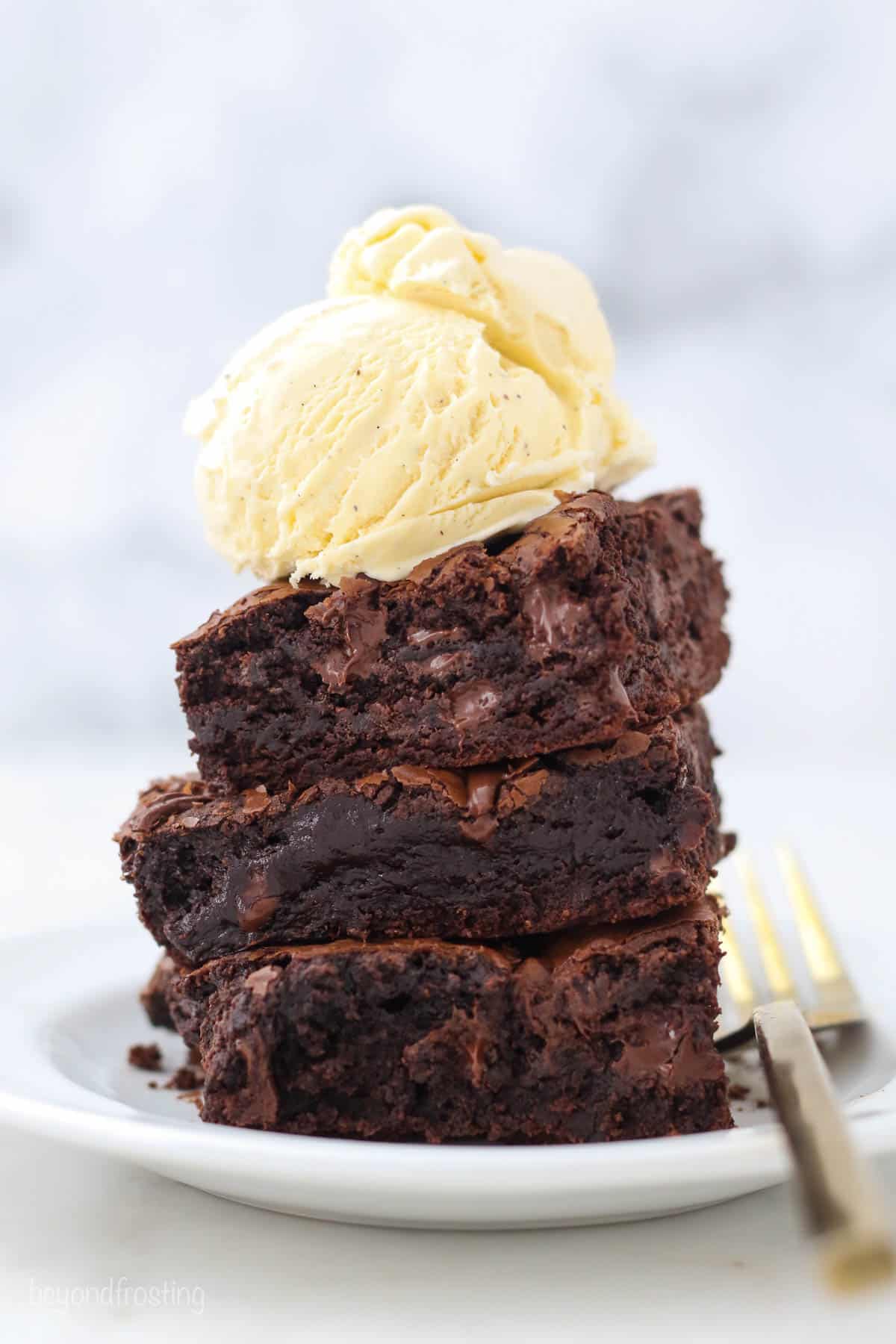 Three brownies on a white plate with a big scoop of vanilla ice cream on top and gold fork laying on the plate