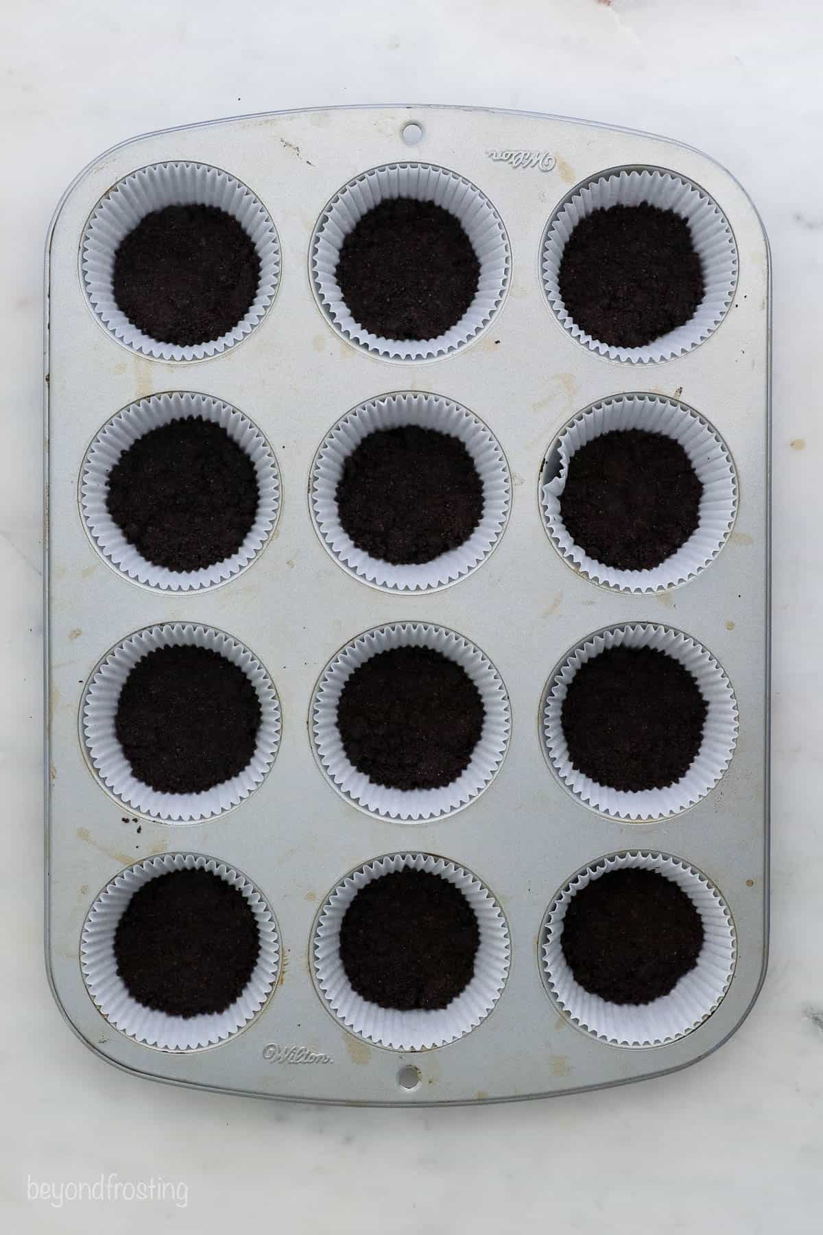A cupcake tin fitted with liners that are filled with Oreo cookie crust