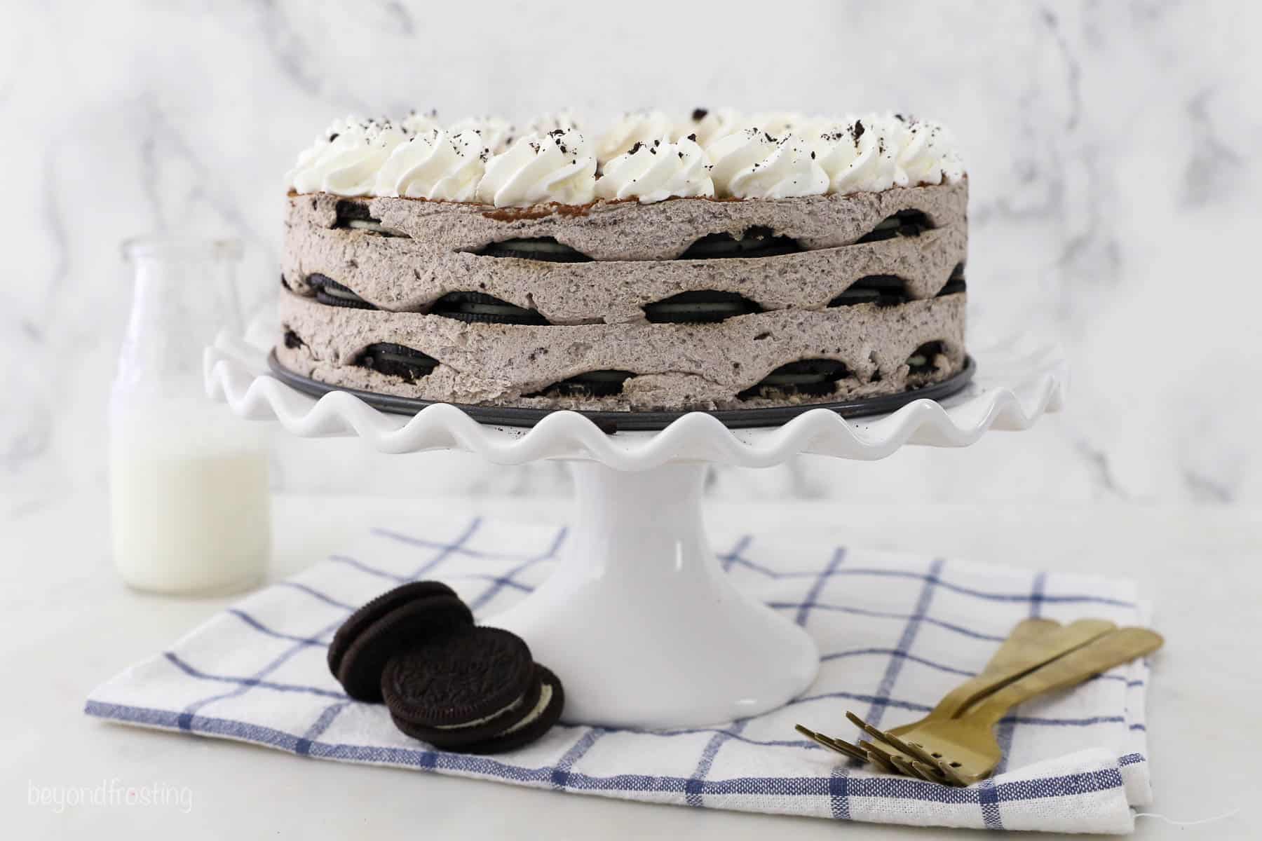 A white ruffled cake stand with a full Oreo mousse cake, three placed forks and 3 Oreos