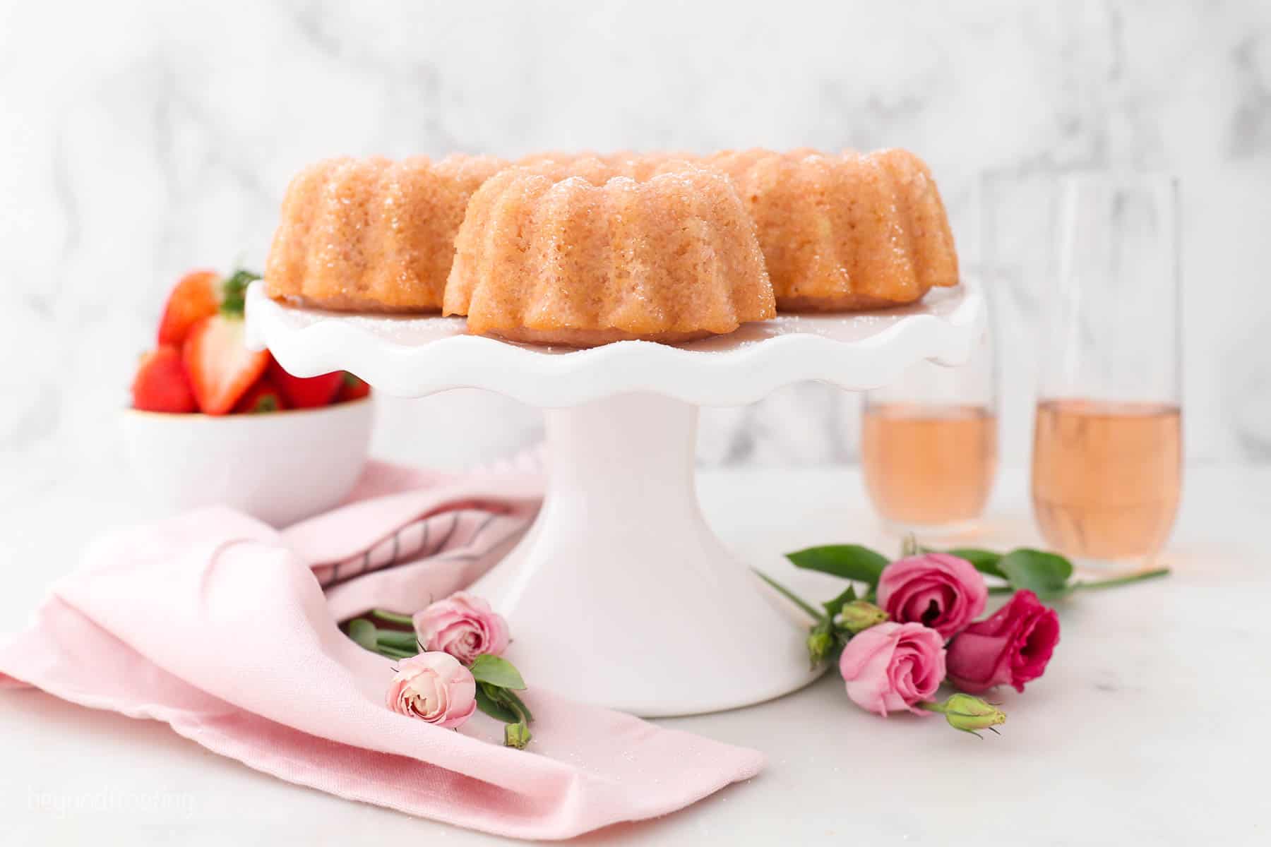 A white cake stand sitting on a pink napkin with mini roses. 4 mini Pink champagne bundt cakes on top