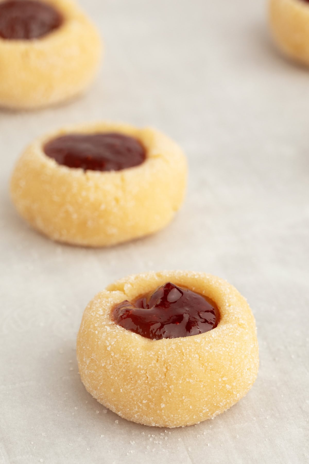 Thumbprint cookies are buttery, melt-in-your-mouth shortbread cookies rolled in sugar, filled with fruit jam, chocolate, caramel, and more. Baked thumbprint cookies filled with jam on a sheet of parchment paper.
