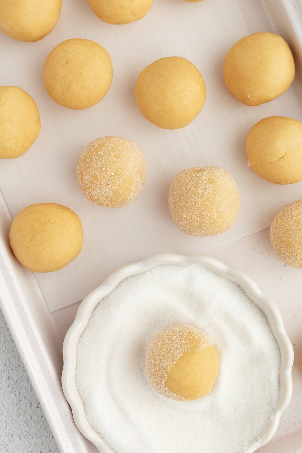 Thumbprint cookies are buttery, melt-in-your-mouth shortbread cookies rolled in sugar, filled with fruit jam, chocolate, caramel, and more. Overhead view of a cookie dough ball in a bowl of sugar, next to more cookie dough balls on a baking sheet.