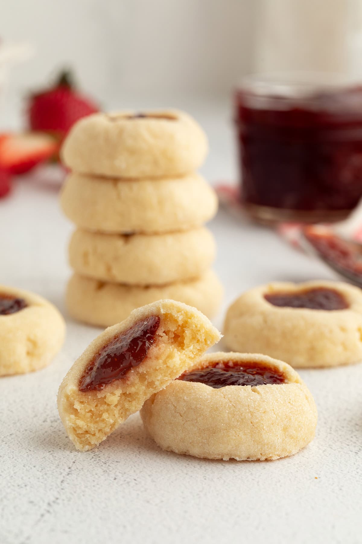 Thumbprint cookies are buttery, melt-in-your-mouth shortbread cookies rolled in sugar, filled with fruit jam, chocolate, caramel, and more. Two jam-filled thumbprint cookies in front of a stack of cookies on a countertop.