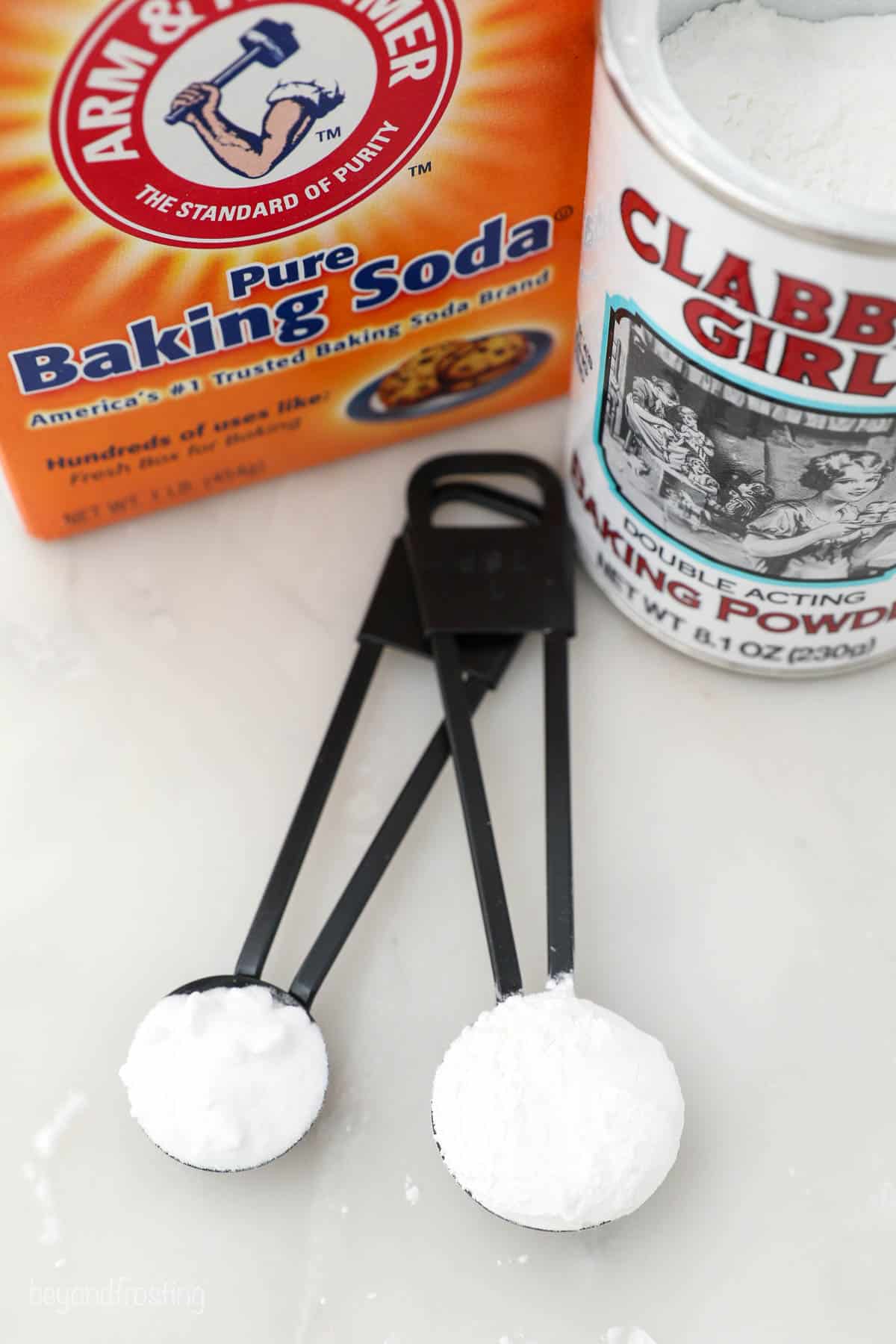 Baking soda in a measuring spoon beside baking powder in a larger measuring spoon on a countertop