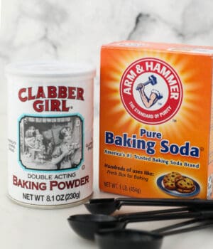 A tub of baking powder beside a box of baking soda with a set of measuring spoons in the foreground
