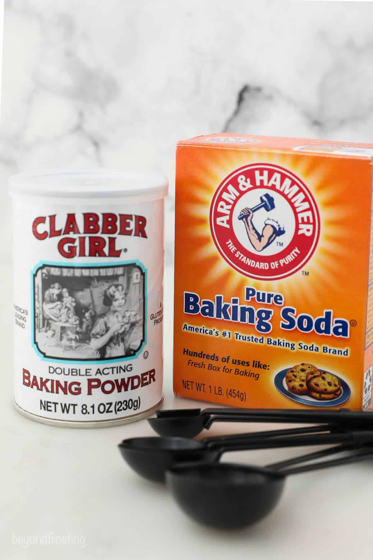 A tub of baking powder beside a box of baking soda with a set of measuring spoons in the foreground