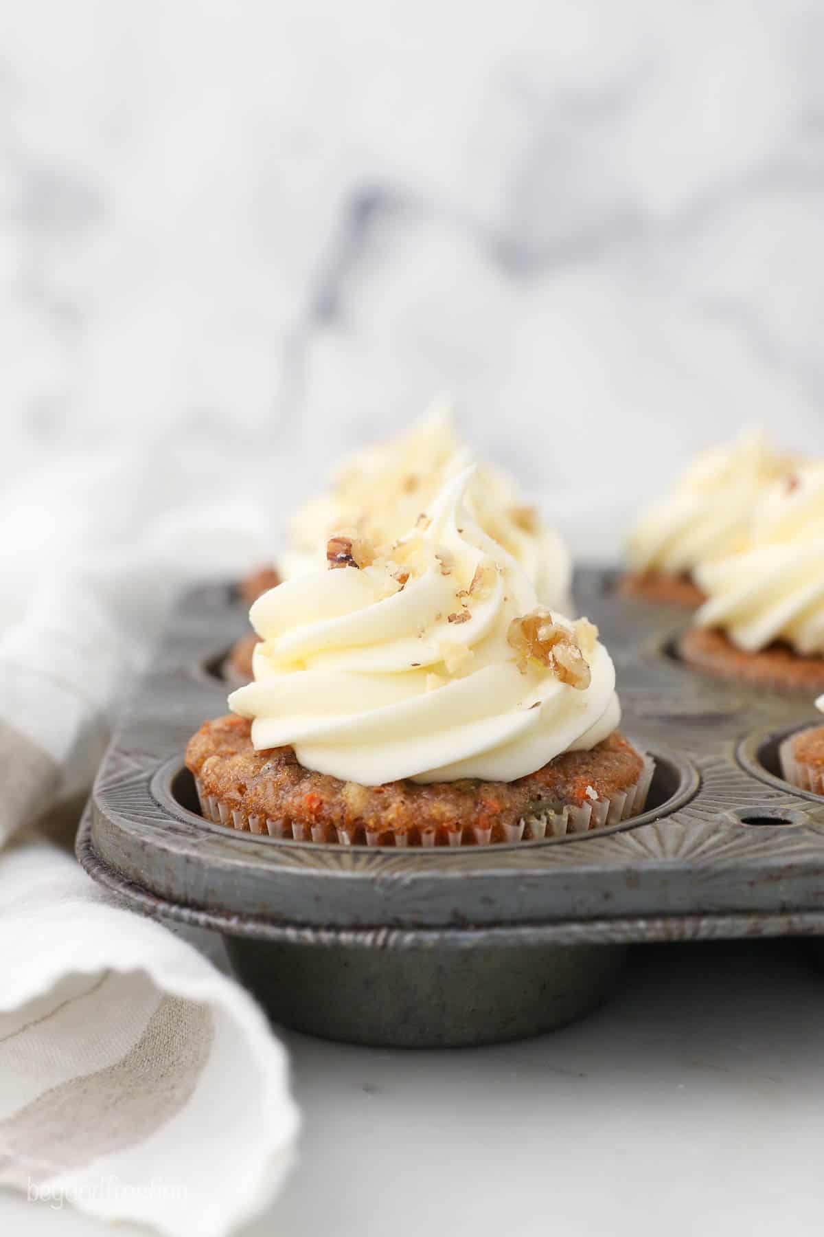 Freshly-baked cupcakes inside of a metal muffin tin with a cloth napkin leaning up against it