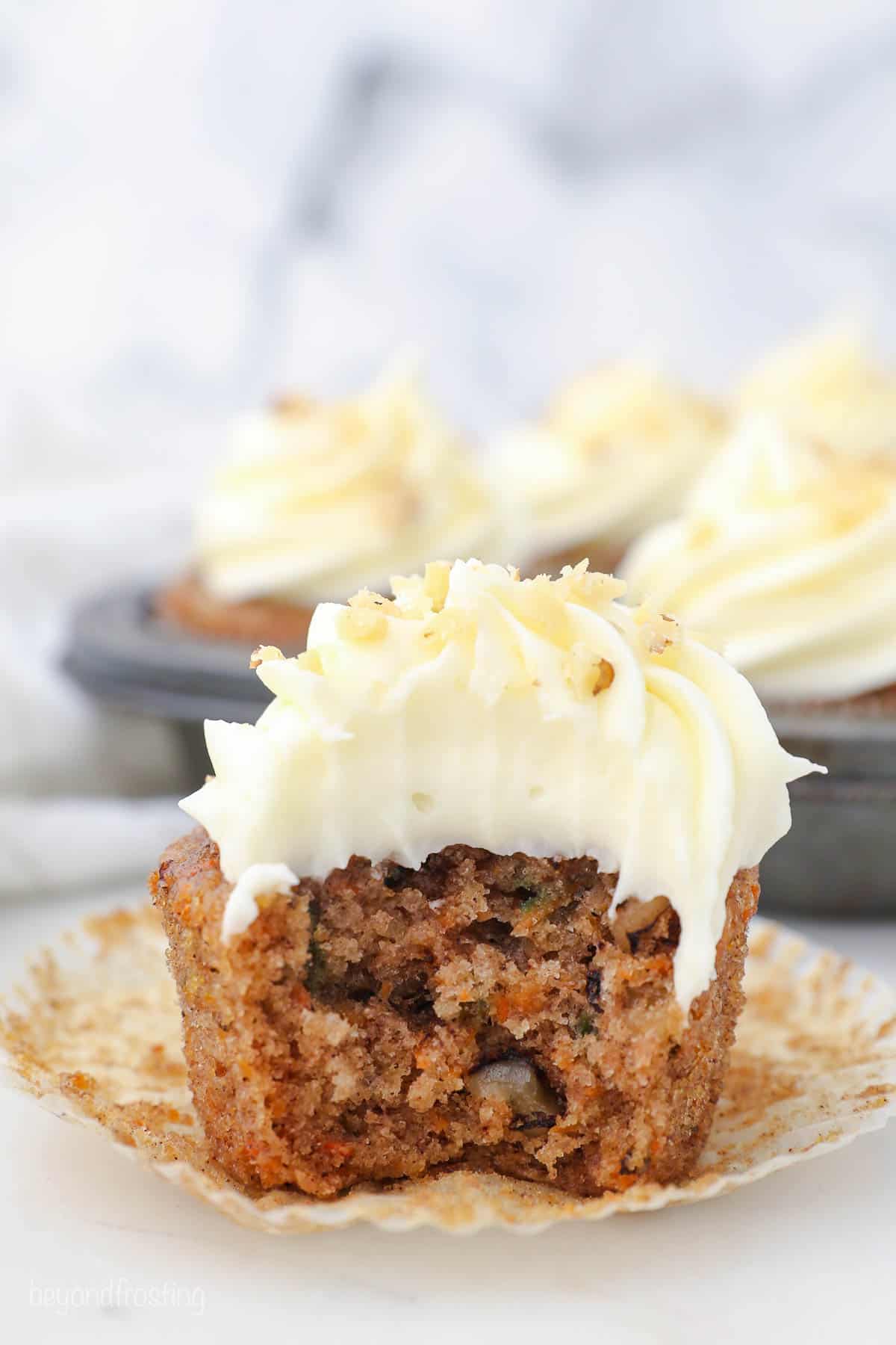 A carrot cake cupcake with a bite taken out of it and a pan containing more cupcakes behind it