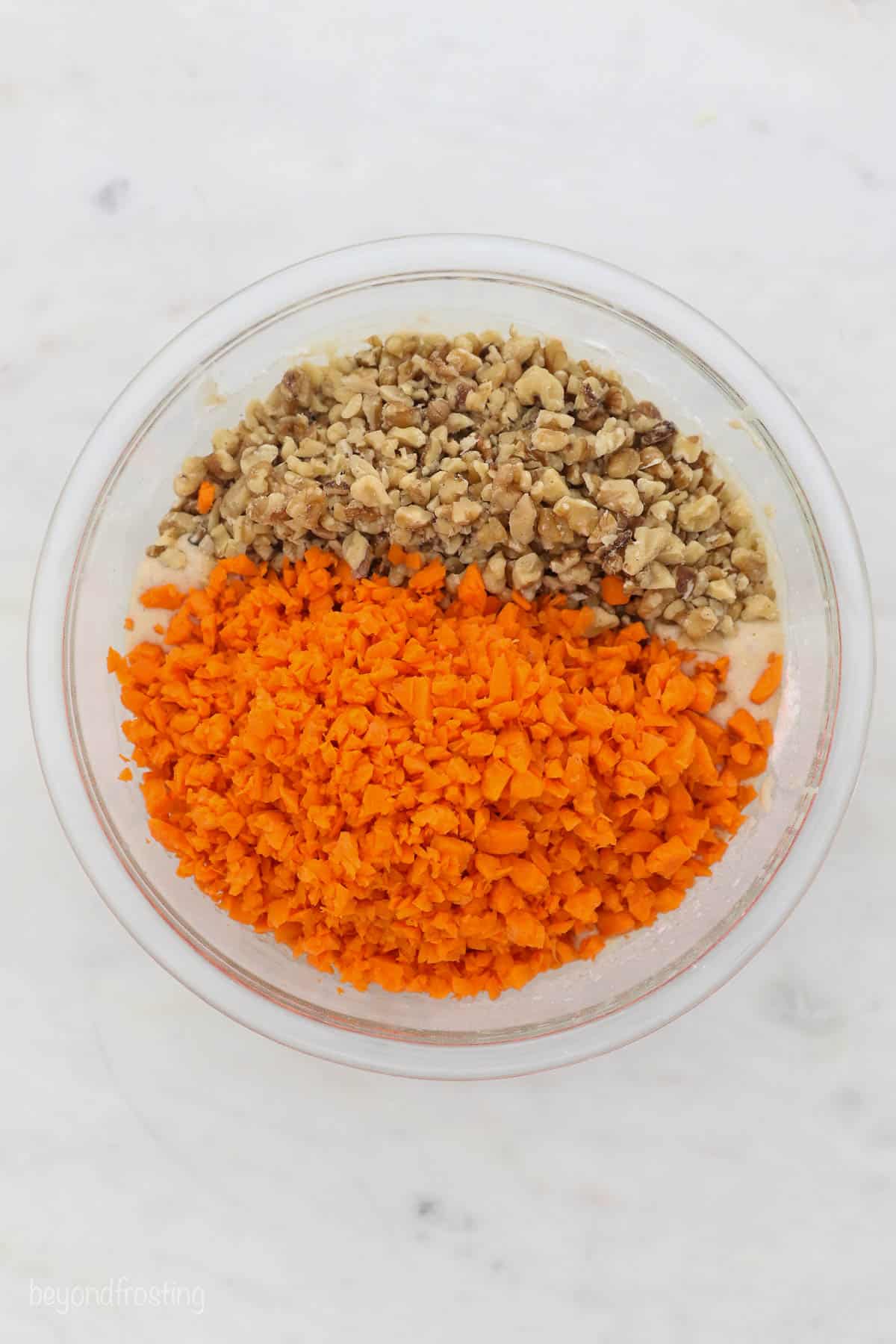 A glass mixing bowl with carrot cake batter, chopped carrots and walnuts