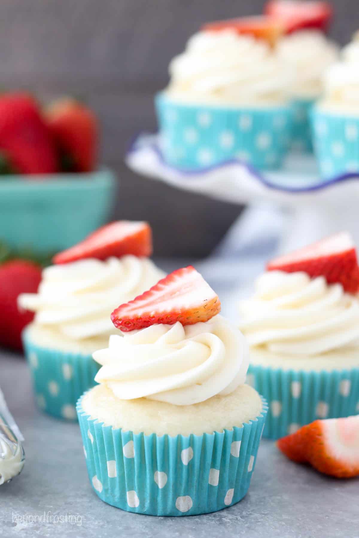 My cream cheese frosting recipe starts with cold ingredients for a thick, velvety frosting that's perfect for decorating cakes and cupcakes. Three vanilla cupcakes topped with swirls of cream cheese frosting and strawberry halves.