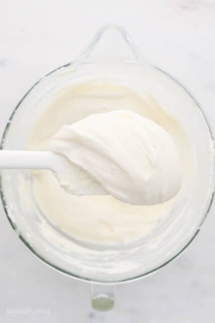 My cream cheese frosting recipe starts with cold ingredients for a thick, velvety frosting that's perfect for decorating cakes and cupcakes. A white spatula with cream cheese frosting being held over a glass mixing bowl.