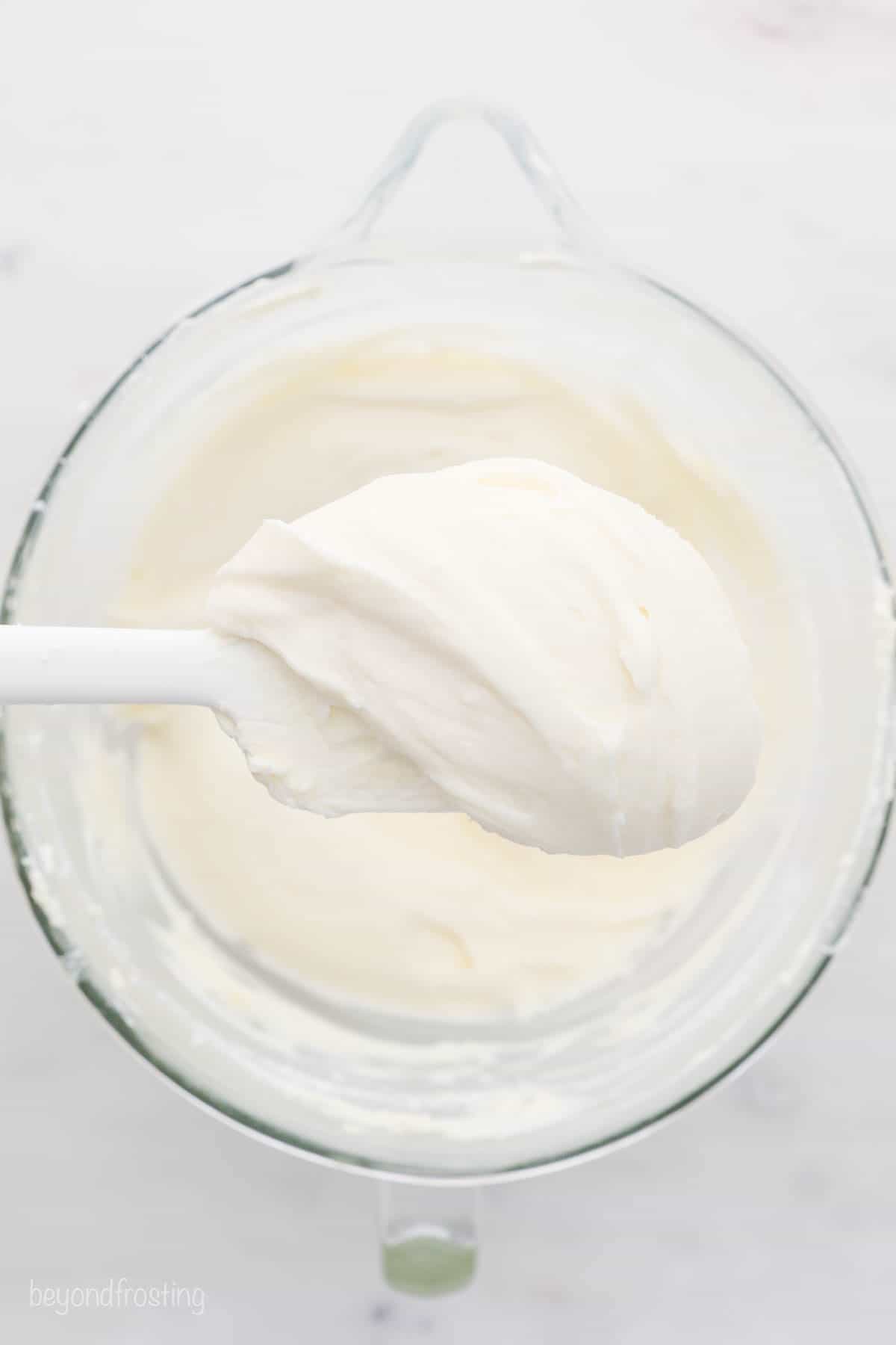A white spatula with cream cheese frosting being held over a glass mixing bowl