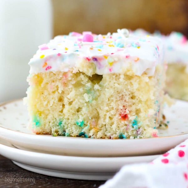 A closeup of a slice of funfetti poke cake with sprinkles on two stacked white plates.
