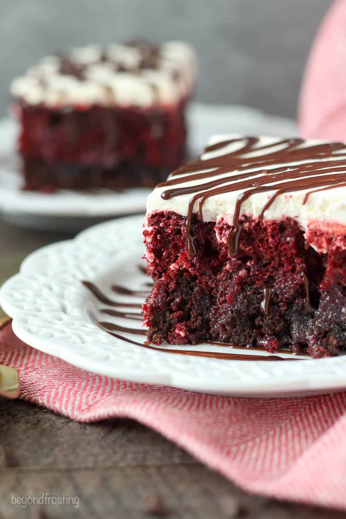A slice of red velvet brownie cake on a plate with another slice on a dish behind it