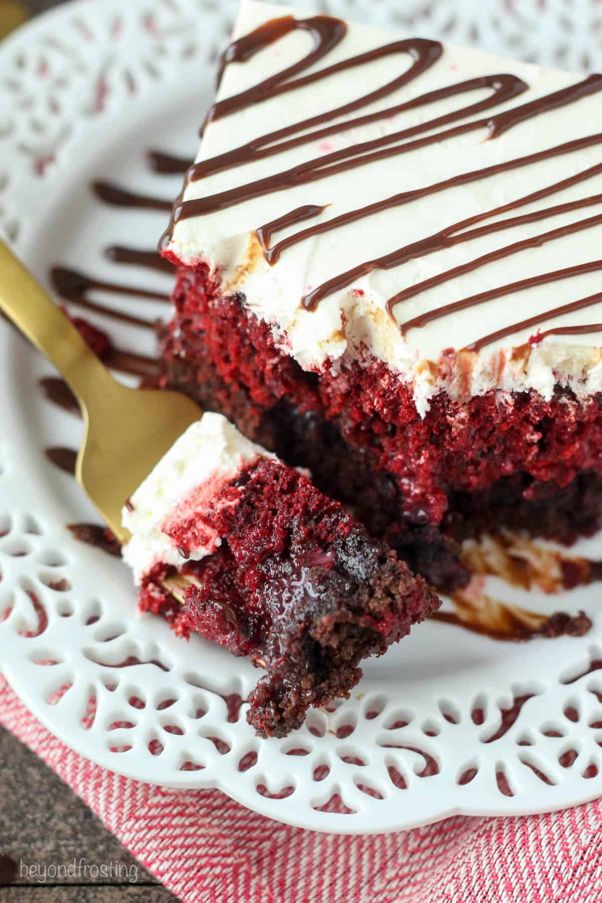 A slice of frosted red velvet brownie cake on a plate with one bite on a golden fork