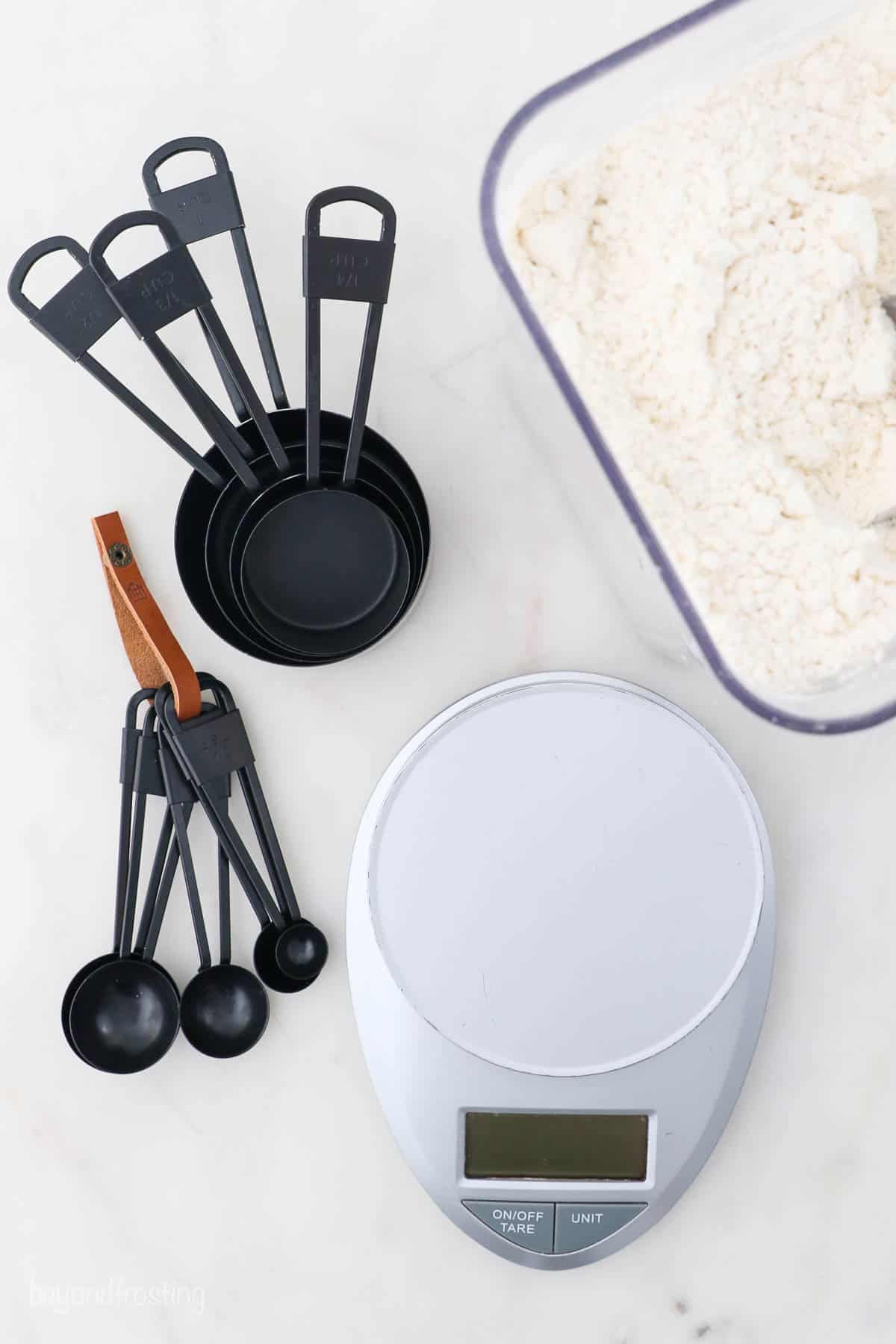 A food scale, a tub of flour, measuring spoons and measuring cups arranged on a marble surface