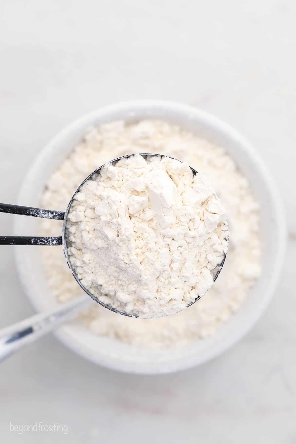 A measuring cup overflowing with flour that has been spooned into it little by little