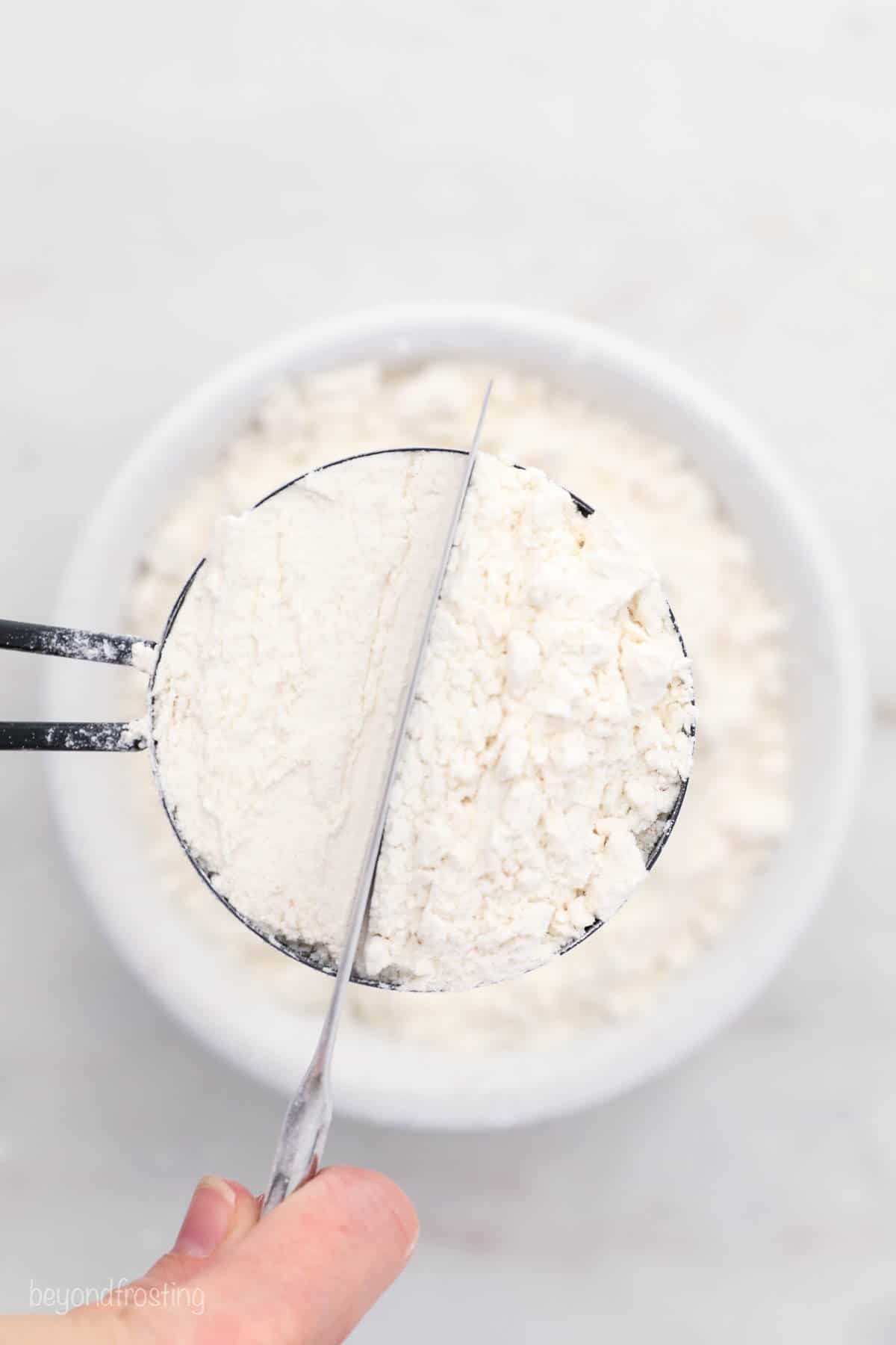 A butter knife being used to level off a cup overflowing with flour at the top