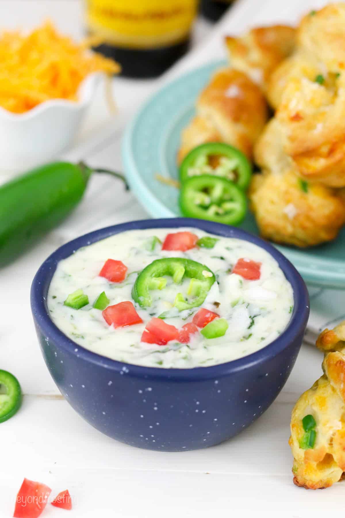 bowl of cheddar jalapeño dip garnished with tomato and jalapeño with a plate of soft pretzels in the background