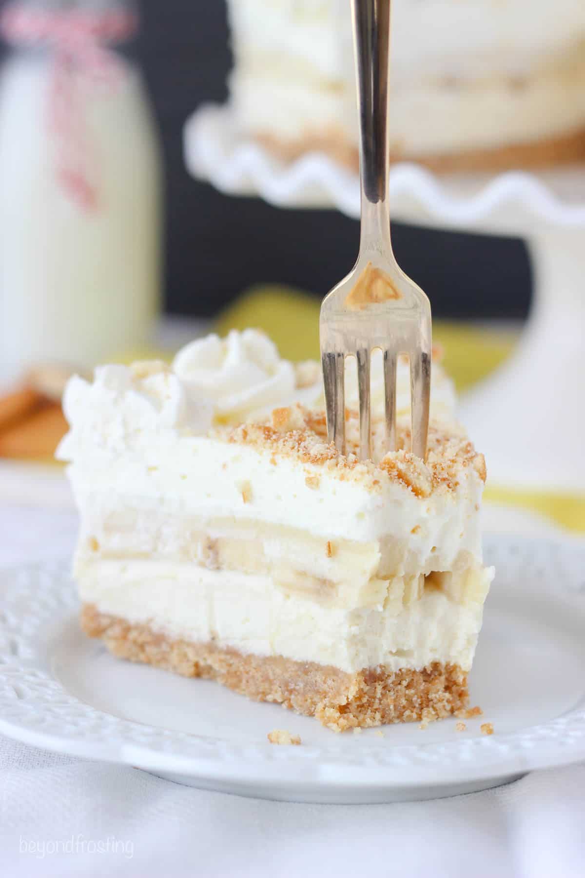 A slice of banana pudding cheesecake on a plate with a couple of bites missing and a fork sticking into it
