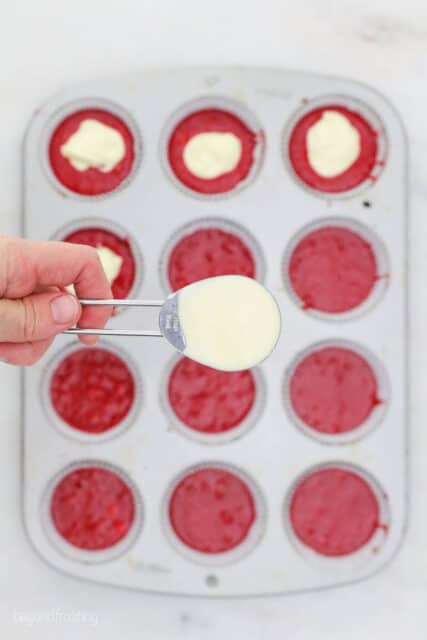 A tablespoons of cream cheese frilling being held over a cupcake pan filled with red velvet cake batter