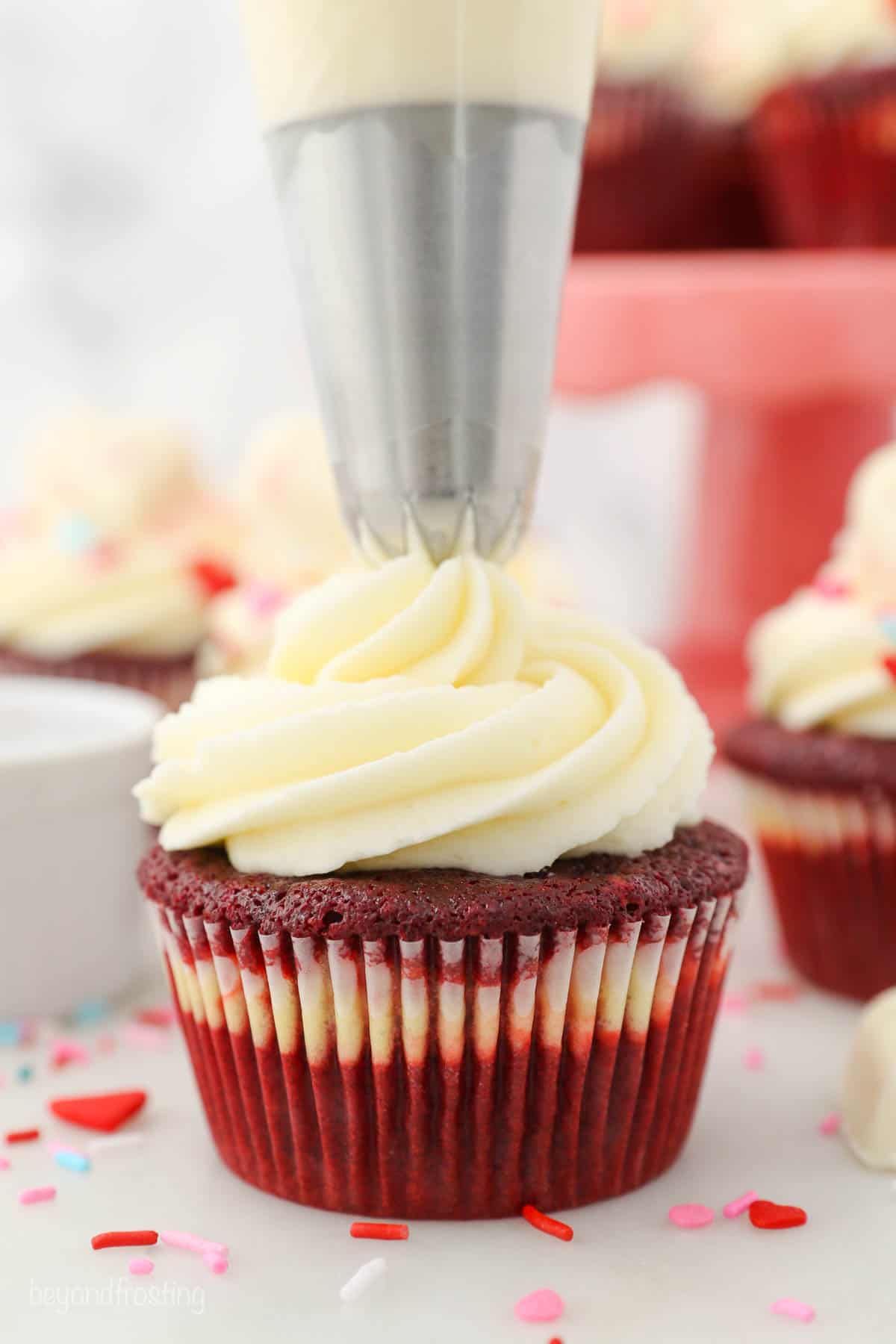A piping bag adding cream cheese frosting on top of a red velvet cupcake
