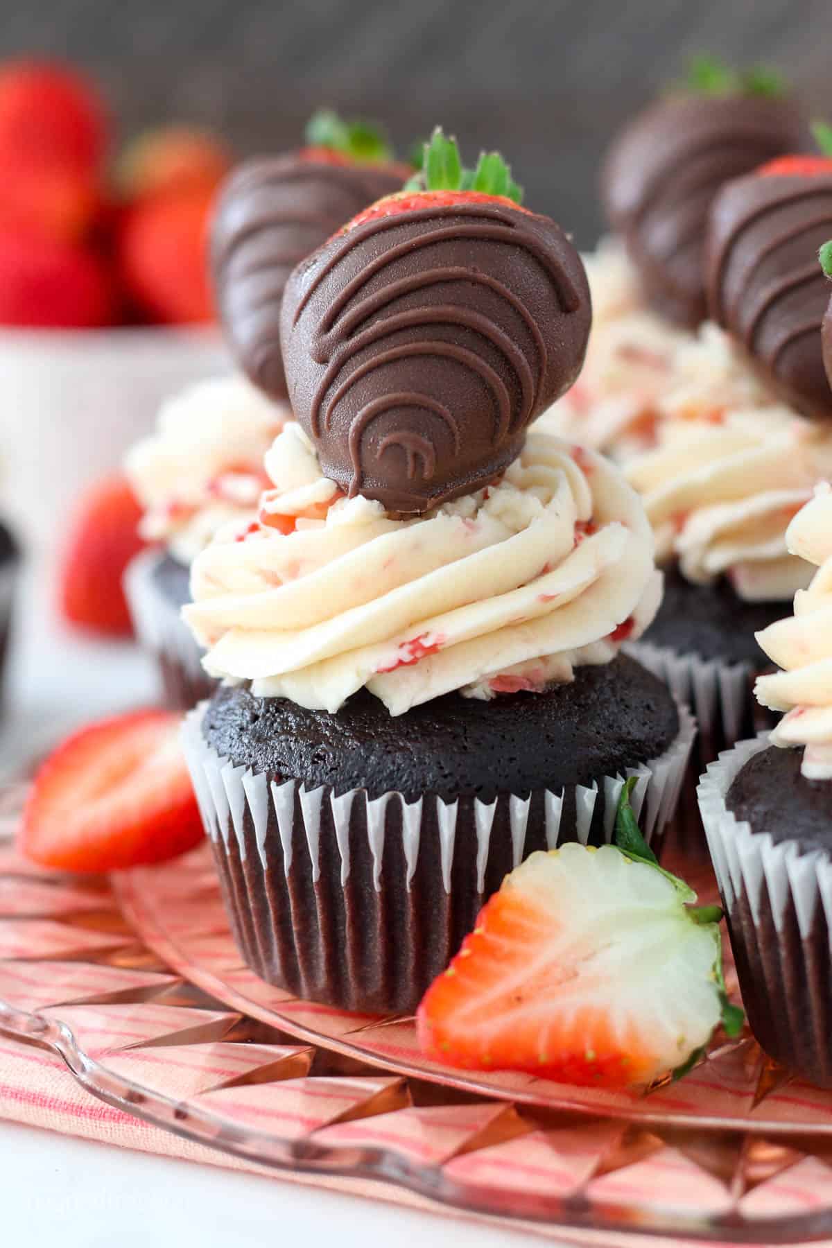 A close up shot of a gorgeous chocolate cupcake with a pretty pink strawberry frosting and a chocolate covered strawberry on top. It's sitting on a pink glass cake plate with a pink striped towel underneath.