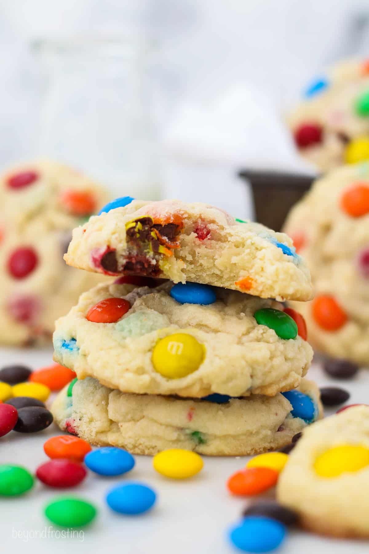 Side view of a stack of three M&M cookies with a bite missing from the top cookie, and more cookies in the background.