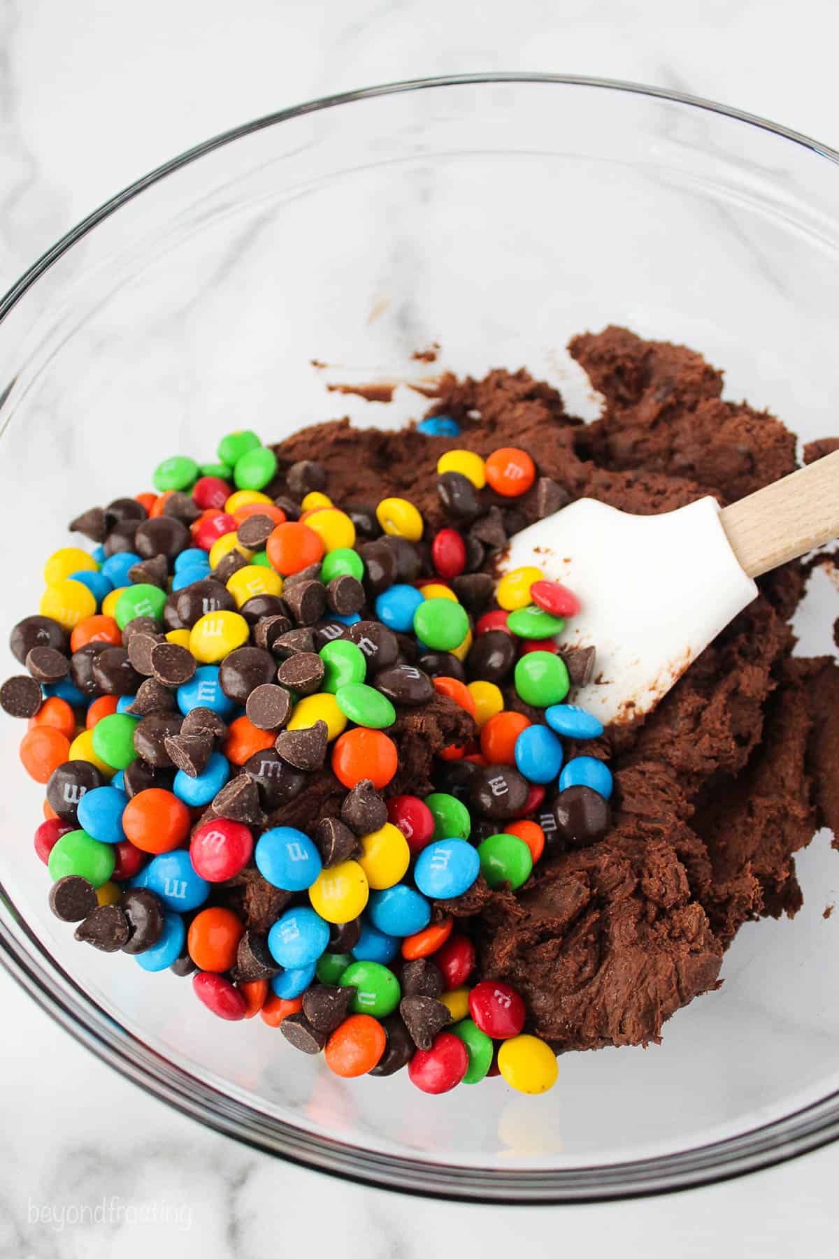 M&Ms added to brownie batter in a glass bowl with a spatula.