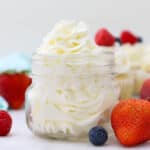 A small glass jar filled with piped whipped cream frosting. The jar is surrounded by berries