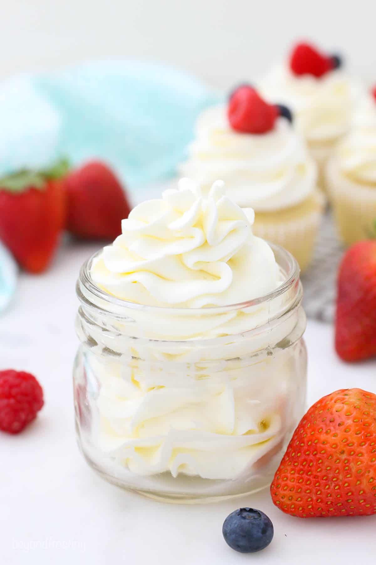 A small glass jar filled with beautifully piped frosting. The jar is surrounded by berries