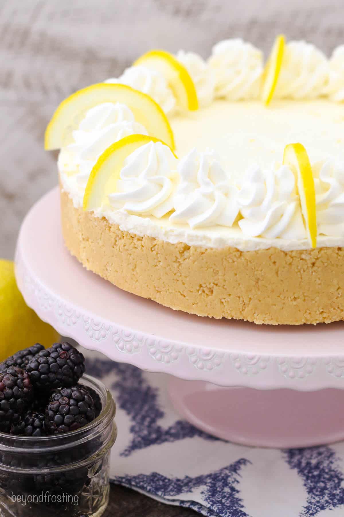 a whole lemon cheesecake on a pink cake stand next to a jar of blackberries