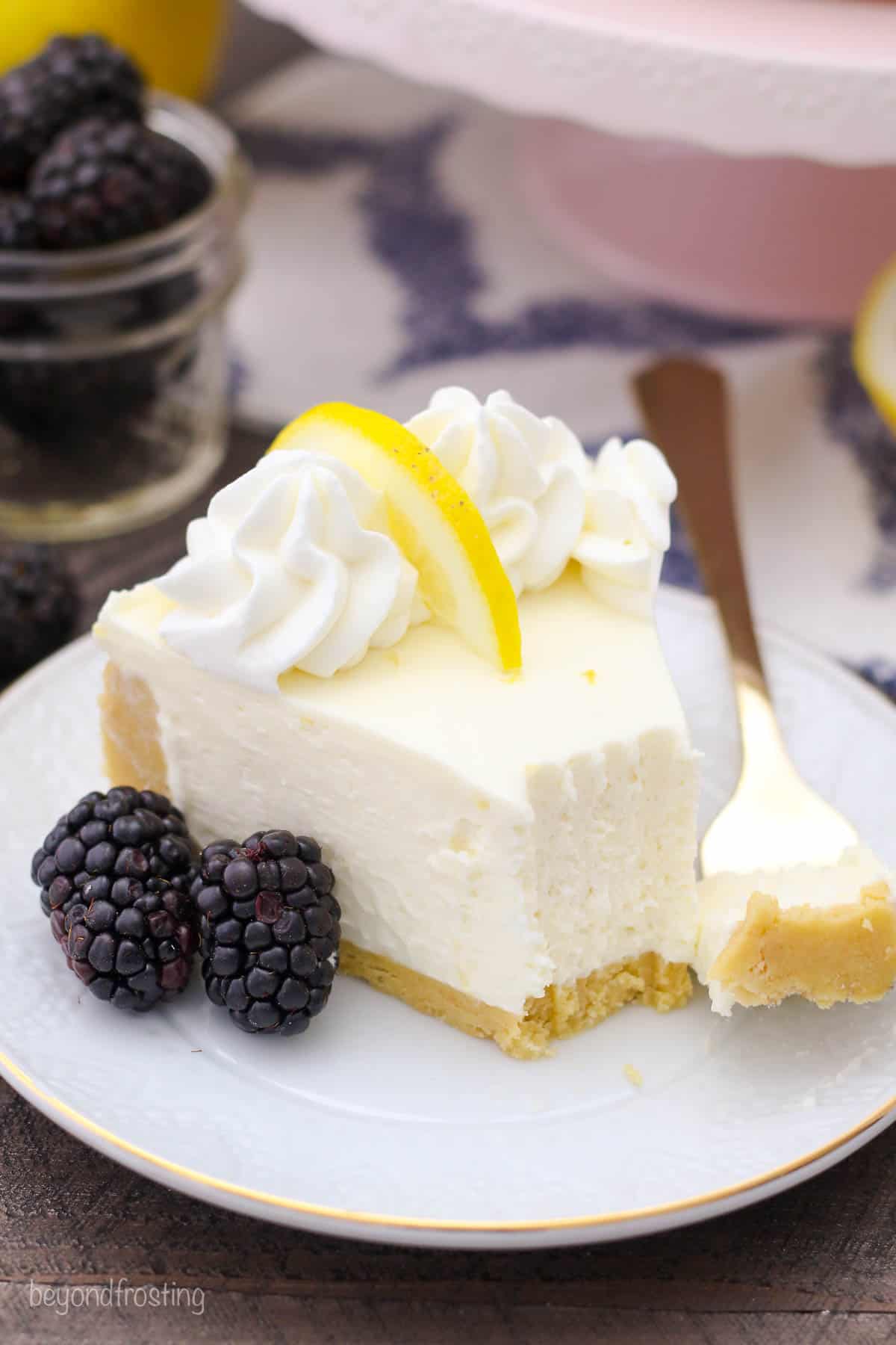 a slice of lemon cheesecake on a gold-rimmed white plate with a bite taken out and blackberries on the side