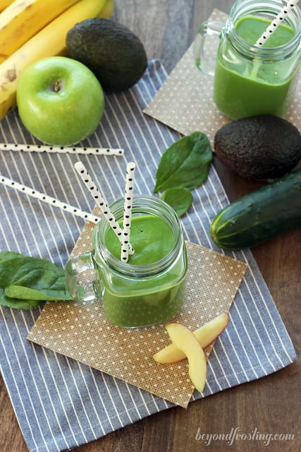 A dining table with two green smoothies sitting on it along with a green apple, avocados and various other produce items