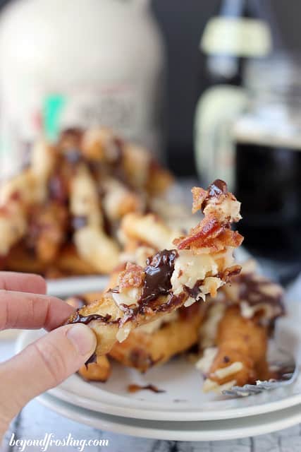 A hand holding a maple bacon donut fry above a plate piled with additional donut fries